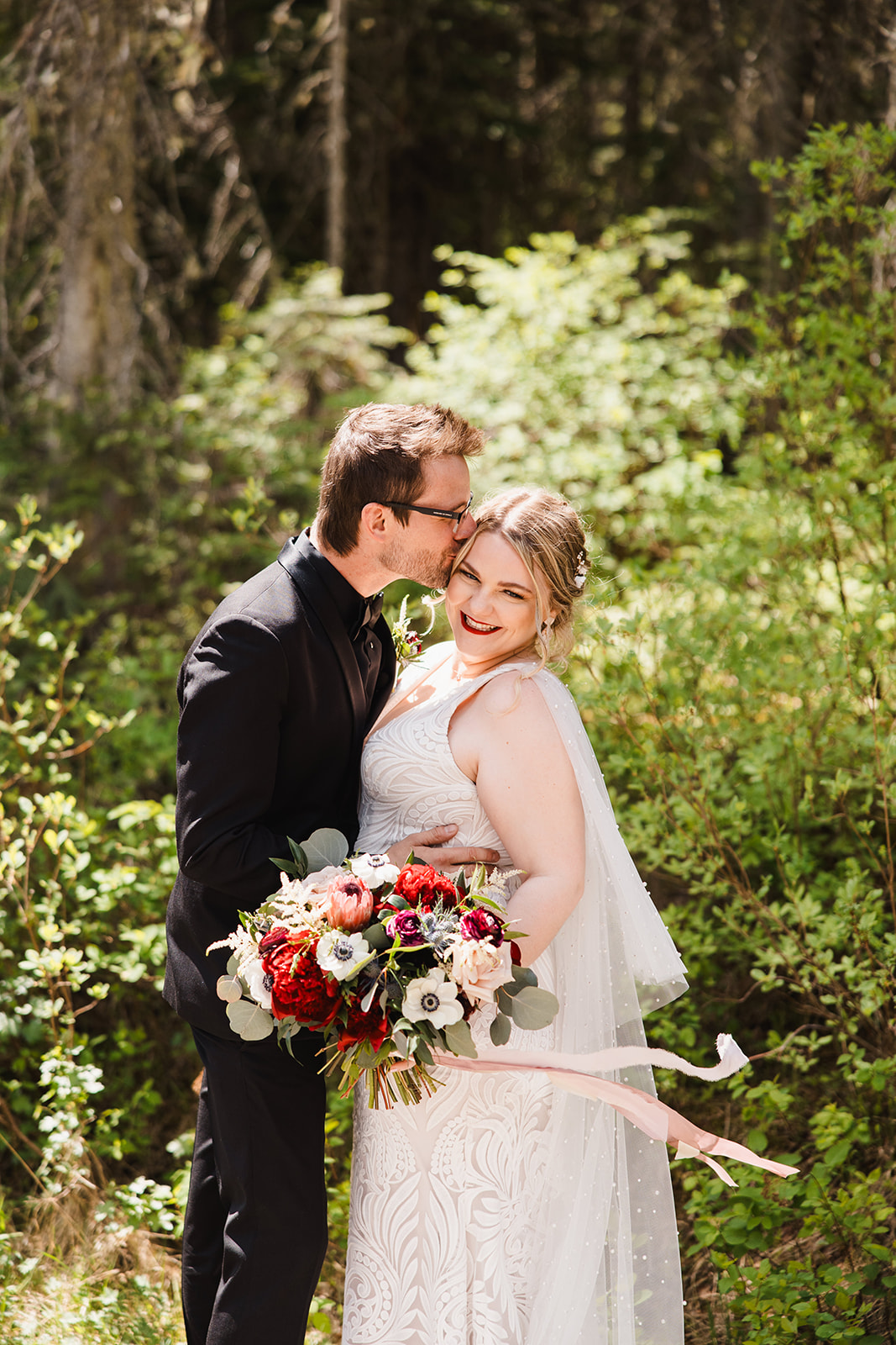 Couple laughing in the forest at Emerald Lake with a red anemone bouquet