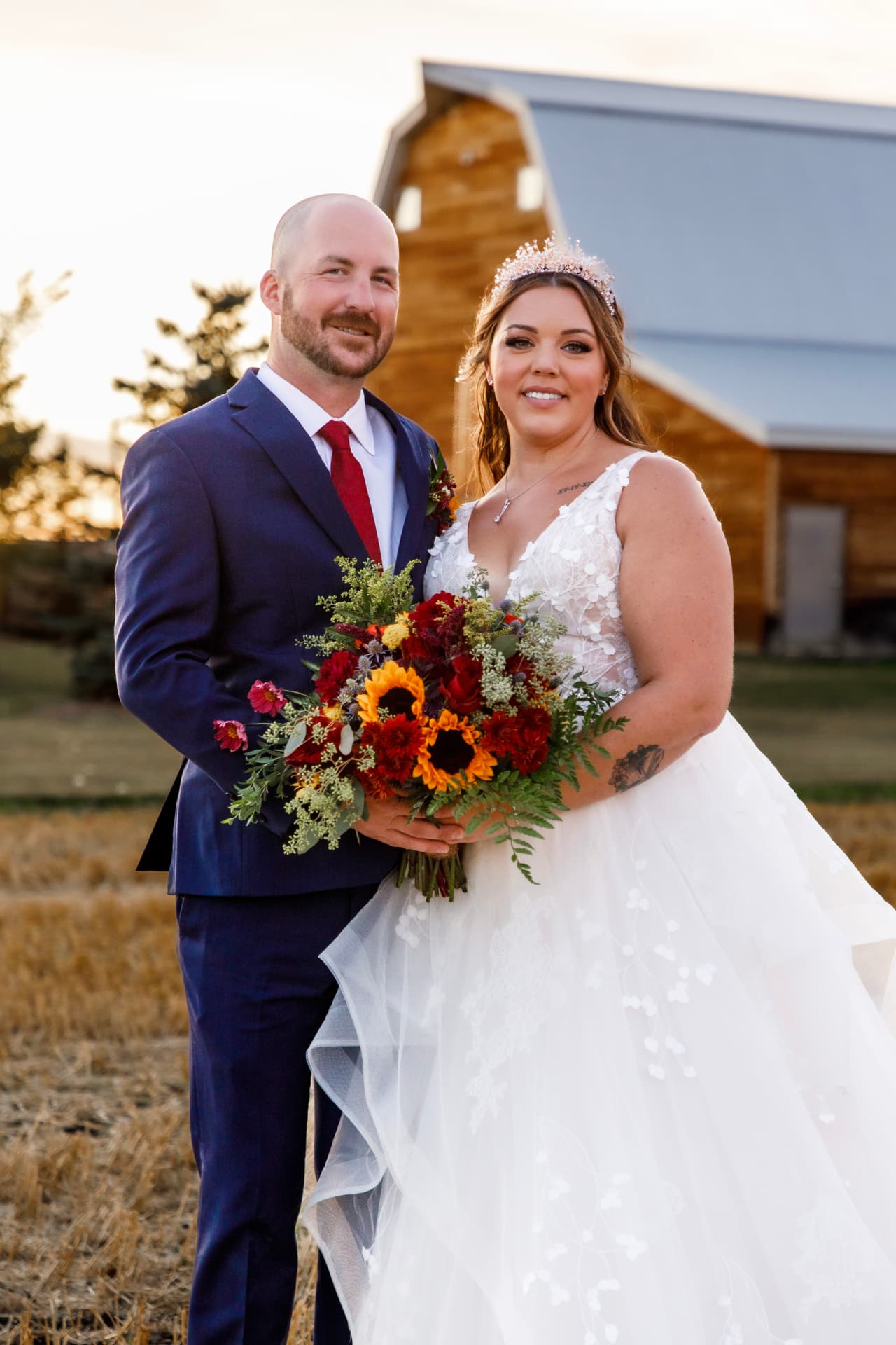 Couple at golden hour in front of a timber barn with a sunflower and dahlia bouquet
